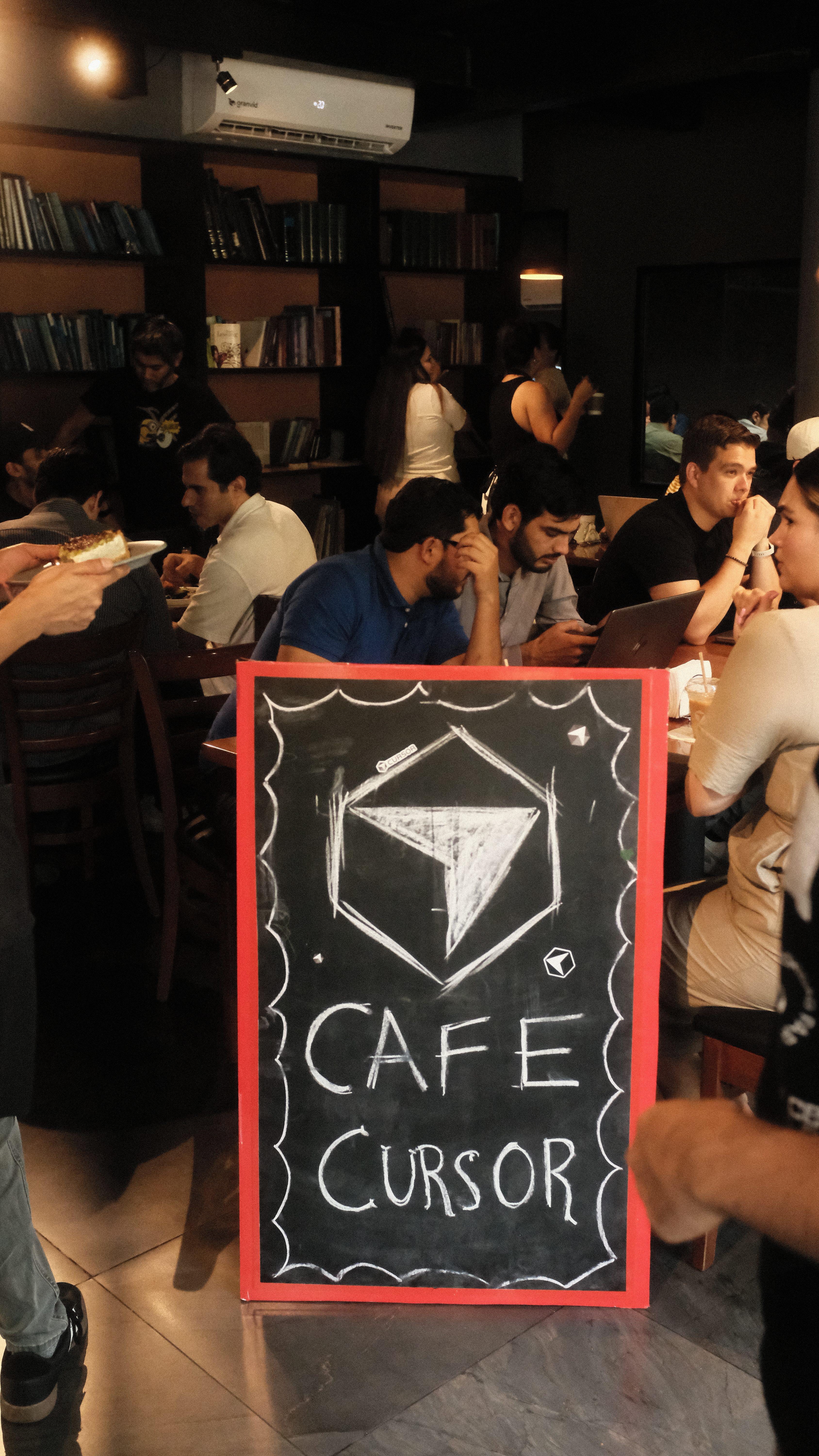 Cafe Cursor chalkboard sign with attendees coding in the background